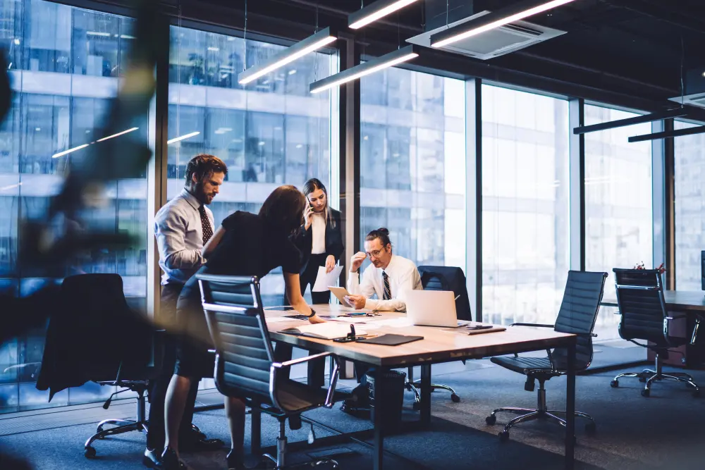 A group of business professionals in a modern office setting collaborating on a project, with large windows showcasing an urban cityscape backdrop.