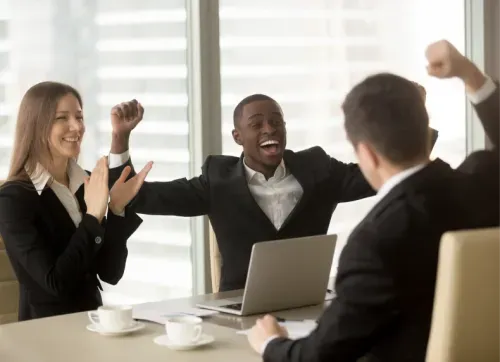 A group of business professionals cheer and celebrate around a table, smiling and raising their hands in excitement after a successful project meeting.