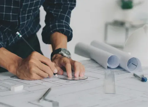 Close-up of an architect’s hands sketching building plans on a blueprint, with drafting tools and rolled-up documents on the table.