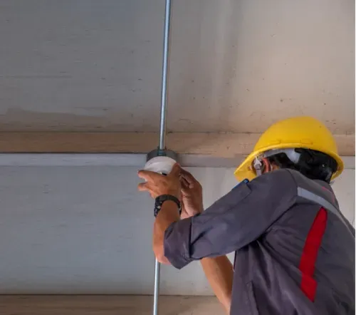 A construction worker wearing a hard hat installs a fire safety system on the ceiling of an office building, ensuring compliance with safety codes.