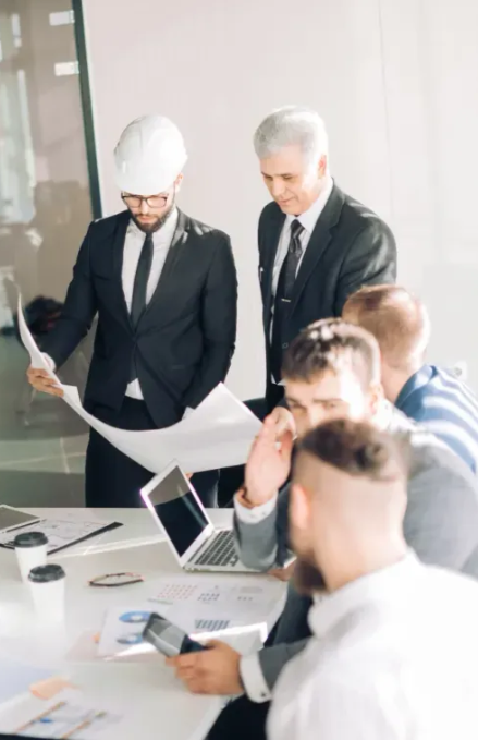 A team of business professionals and an engineer in a hard hat reviewing construction plans during a meeting in a bright corporate office space.