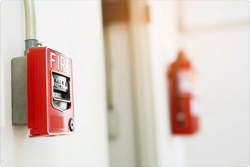 Close-up of a red fire alarm mounted on a white wall with a blurred fire extinguisher in the background, highlighting fire safety equipment.
