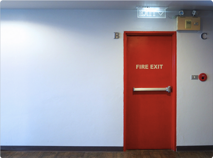 A red fire exit door in a hallway with an illuminated exit sign above it, emphasizing emergency escape route. The wall is plain white.