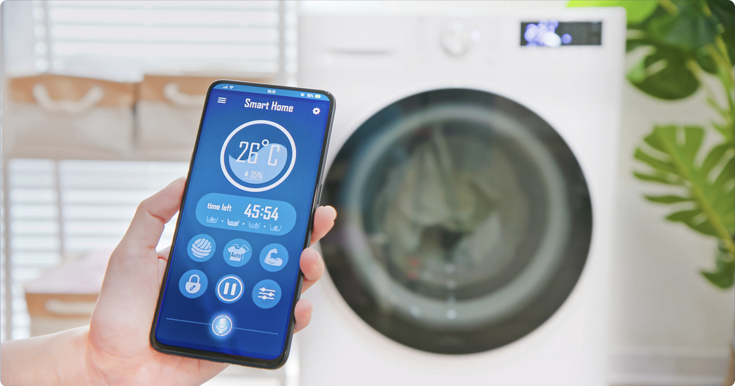 Hand holding a smartphone with a smart home app controlling a washing machine in the background, showing temperature and remaining time."