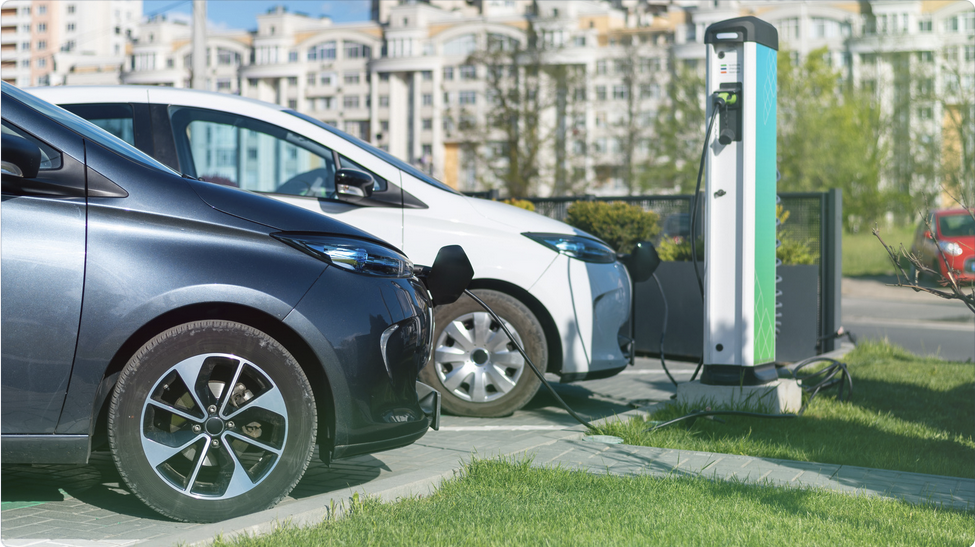 Two electric cars plugged in and charging at a public charging station in an urban setting with buildings in the background.