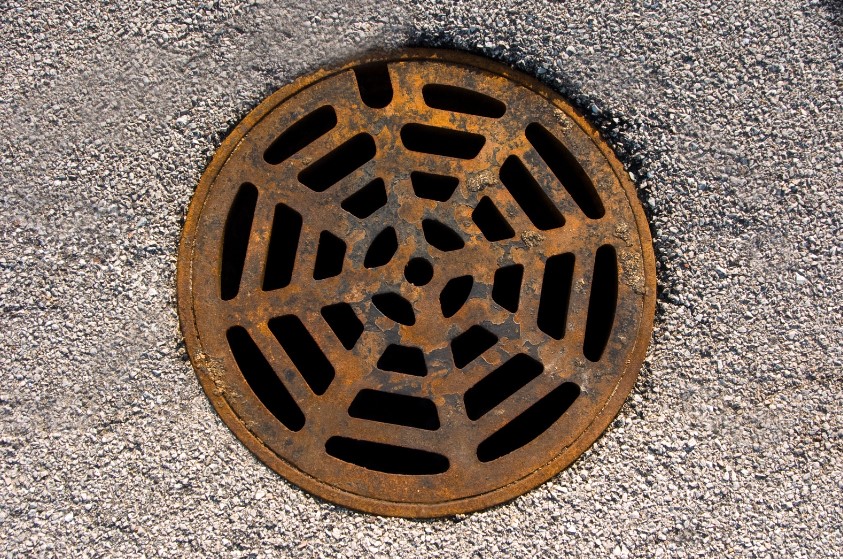 Close-up view of a rusty metal drain cover with a unique, circular design pattern, set against a rough, textured surface of asphalt.