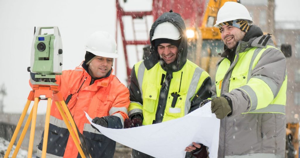 Three construction workers wearing safety gear and hard hats examine blueprints next to surveying equipment in a snowy outdoor setting