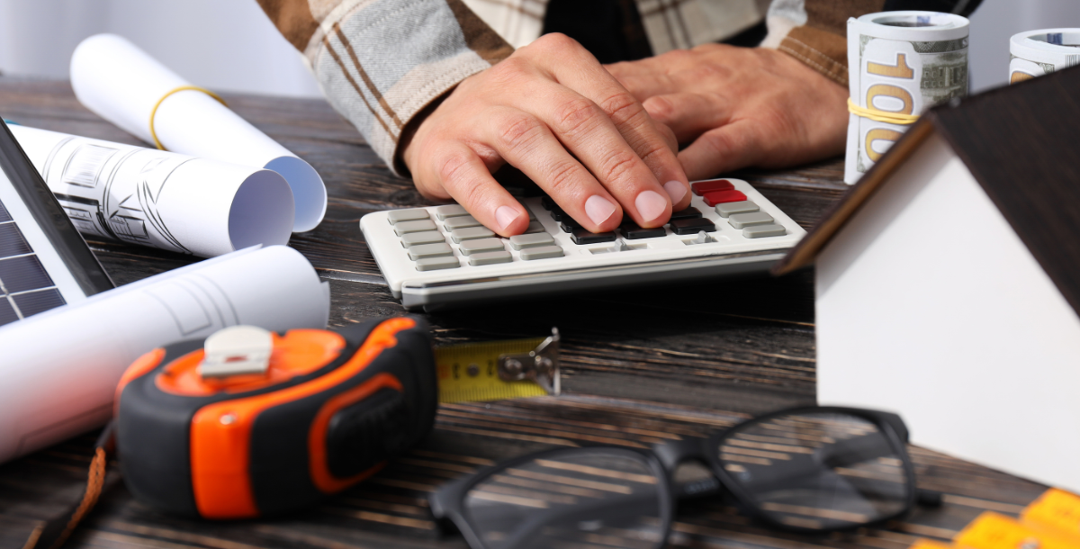 A person in a plaid shirt uses a calculator on a table cluttered with blueprints, rolled cash, a small house model, measuring tape, and construction tools.