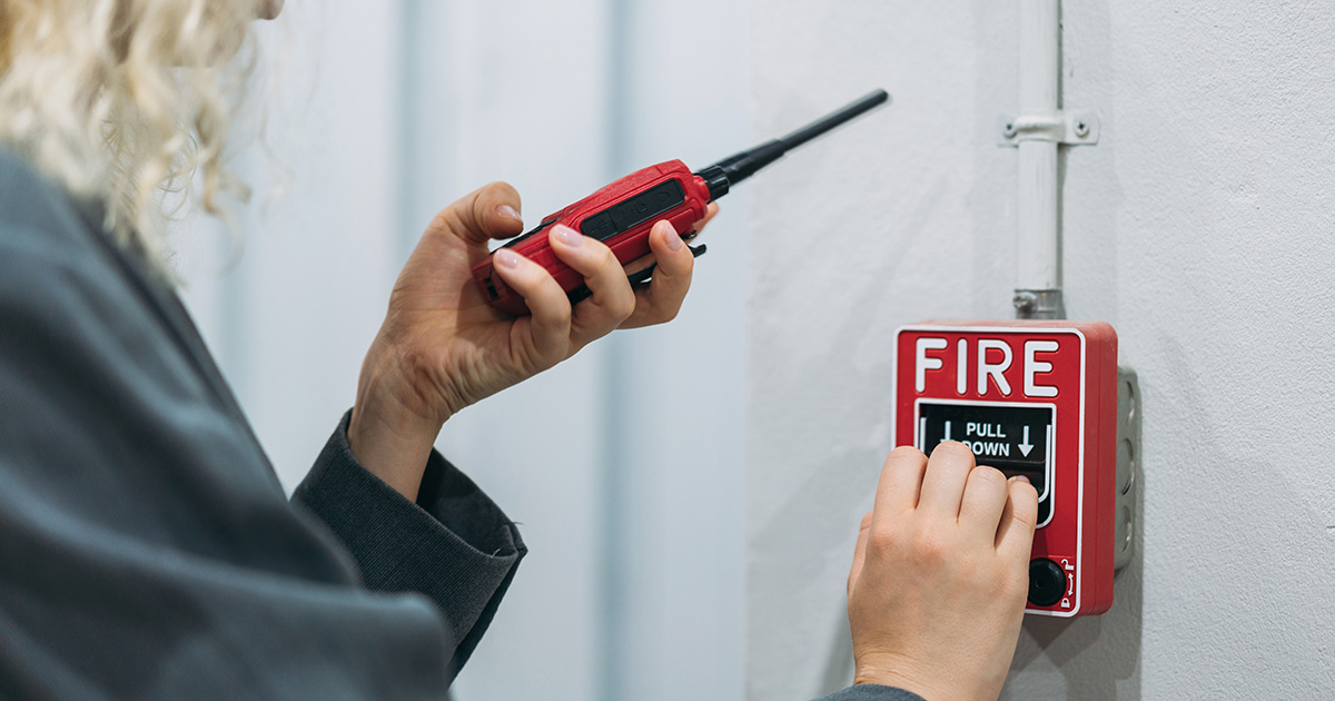 Person in a gray jacket pulls a red fire alarm while holding a walkie-talkie, signaling an emergency. The alarm is mounted on a white wall with wiring above.
